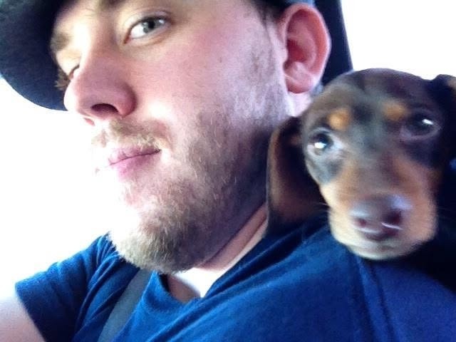 Wes driving with Boo the dachshund riding on his shoulder, both looking at the camera